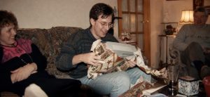 Adult retro gamer opening a wrapped gift with family in a 1990s living room